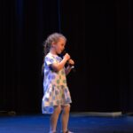 Dziewczynka na scenie śpiewa trzymając mikrofon z tyłu slajd OŚRODEK KULTURY w Sejnach XXI FESTIWAL TAŃCA |PIOSENKI "O ZŁOT PANTOFELKI SKOWRONKA"