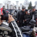 dłonie trzymające telefon robiąc zdjęcie dla kobiety i dwojga mężczyzn w czapkach z daszkiem na tle motocykli i dużej grupy ludzi