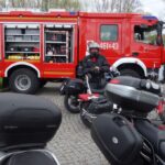trzy motocykle z przodu za nimi mężczyzna w kasku a za nim stoi wóz strażacki i dwóch strażaków przy drzwiach
