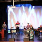 sześciu mężczyzn na scenie grają na instrumentach: pianino, trąbka, saksofon, perkusja, gitara, tara, przed nimi stoi kobieta i śpiewa