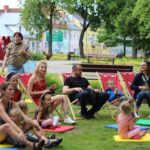 11 osób kobiety dzieci i mężczyzna w parku siedzą na trawie i leżaczkach
