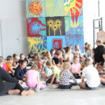 25 dzieci siedzi na podłodze. Na ścianie wiszą kolorowe plansze