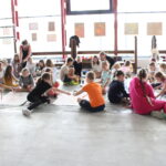 25 dzieci siedzi na podłodze i malują, nad nimi stoi instruktorka.
