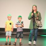 dziewczyna z torba na ramieniu trzyma mikrofon stojąc na scenie obok niej dwaj mali chłopcy