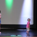 dziewczynka stoi na scenie z mikrofonem obok niej na scenie biega dziecko