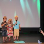 dwóch chłopców i dziewczynka stoją na scenie z mikrofonem