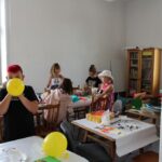 4 dziewczynek przy stole wyklejaja z bibuły z przodu chłopiec dmucha żółty balon