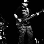 czarno - białe zdjęcie na którym mężczyzna stoi przed mikrofonem z gitarą na scenie