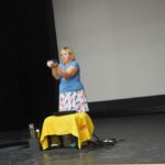 kobieta na scenie za żółtym stolikiem trzyma w dłoni bańki mydlane