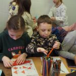 dzieci siedzą i stoją kolorują i składają origami