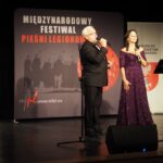 kobieta i mężczyzna stoją na scenie i śpiewają, za nimi napis Międzynarodowy Festiwal Pieśni Legionowej