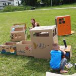 dziecko maluje na kartonie za nim siedzi kobieta na trawie