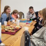 dzieci i dorośli siedza przy stole grają w planszówkę i na stole stoi jedzenie